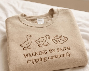 May include: Beige crewneck sweatshirt with embroidered ducks and the text "WALKING BY FAITH tripping constantly". The sweatshirt is folded on a white surface. The brand tag is visible.