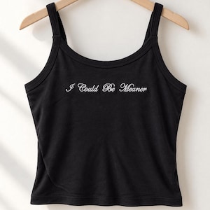 Könnte beinhalten: Ein schwarzes ärmelloses Tanktop mit Rundhalsausschnitt und breiten Trägern. In der Mitte der Brust befindet sich der weiß gestickte Schriftzug "I Could Be Meaner" in einer Schreibschrift. Das Kleidungsstück hängt an einem Holzkleiderbügel.