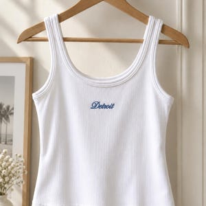 May include: A white ribbed vest top featuring a scoop neckline and thick shoulder straps. The centre of the chest displays the word Detroit embroidered in a blue cursive script font. The garment is displayed on a wooden hanger.