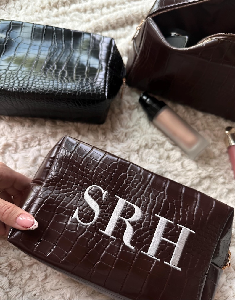 May include: A collection of brown and black faux-leather cosmetic bags with a crocodile skin texture. One bag is embroidered with the initials "SRH" in white. Makeup products are visible in the background.