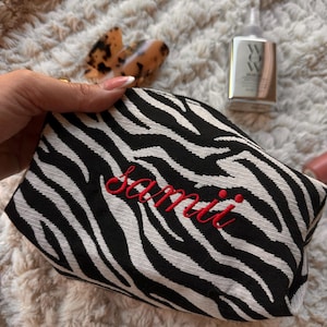 May include: A black and white zebra print cosmetic bag with the name "Sami" embroidered in red. The bag is rectangular and has a zipper closure. Other items in the image include a hair clip and a bottle of lotion.