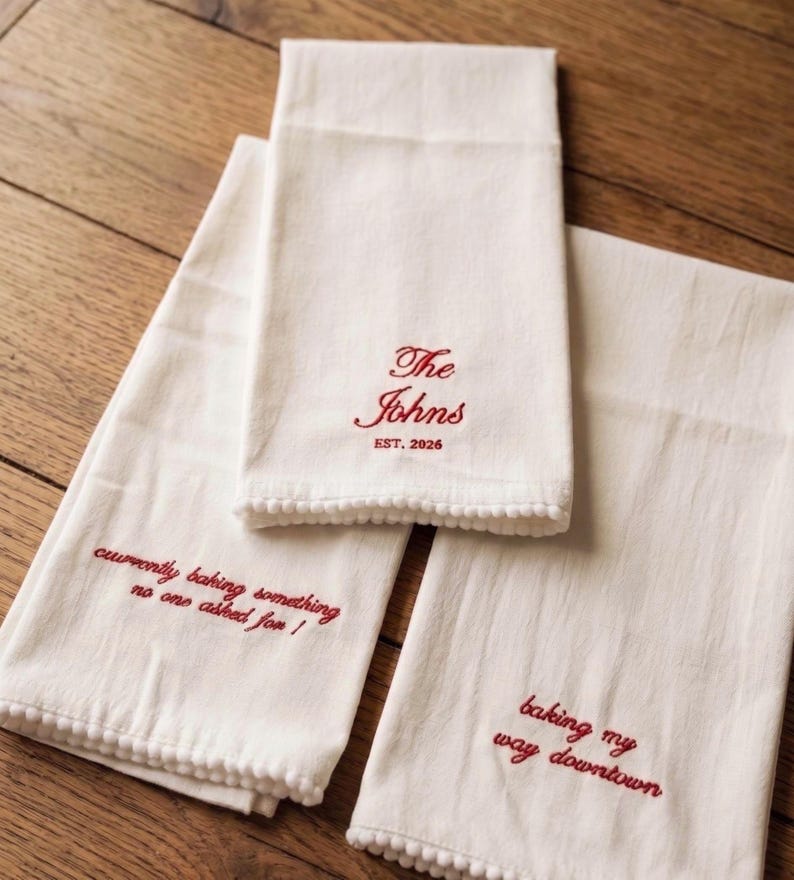 May include: Three off-white kitchen towels with red embroidered text and pom-pom trim. One towel reads The Johns EST. 2026, another says currently baking something no one asked for!, and the third features the text baking my way downtown.