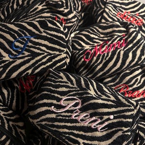 Puede incluir: Una colecci&oacute;n de estuches rectangulares peque&ntilde;os con cremallera y un patr&oacute;n de rayas de cebra en negro y beige. Cada estuche est&aacute; personalizado con nombres bordados en varios colores, como rosa, rojo y azul, en letra cursiva.
