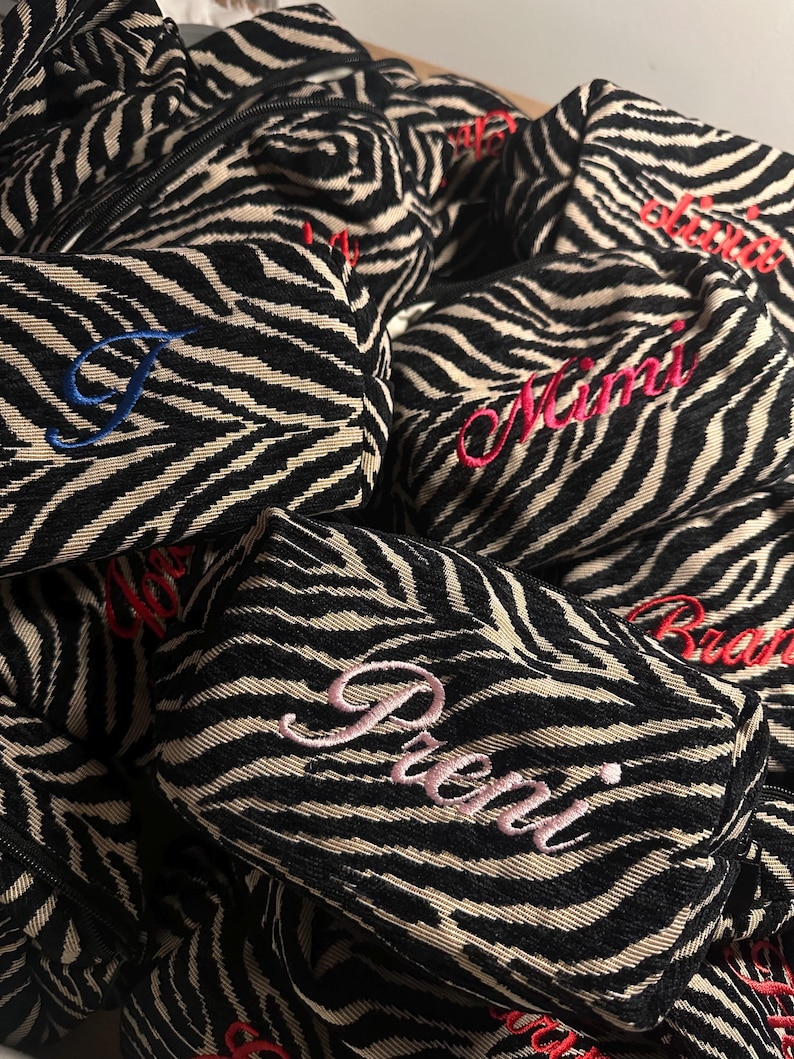 Puede incluir: Una colecci&oacute;n de estuches rectangulares peque&ntilde;os con cremallera y un patr&oacute;n de rayas de cebra en negro y beige. Cada estuche est&aacute; personalizado con nombres bordados en varios colores, como rosa, rojo y azul, en letra cursiva.