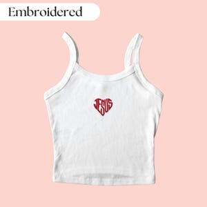 May include: White tank top with red embroidered heart design containing the word "JESUS". The top has thin straps and a scoop neckline. The word "Embroidered" is in a white oval in the upper left corner.