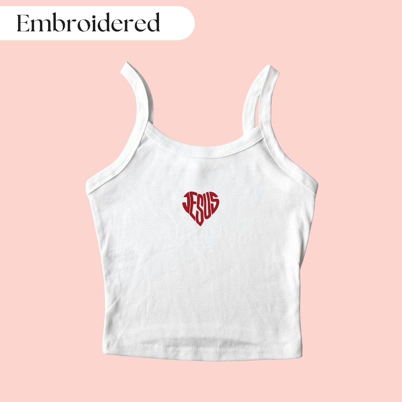 May include: White tank top with red embroidered heart design containing the word "JESUS". The top has thin straps and a scoop neckline. The word "Embroidered" is in a white oval in the upper left corner.