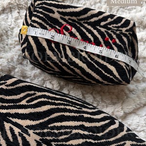 Puede incluir: Un estuche de tama&ntilde;o mediano con estampado de cebra en negro y beige. El estuche presenta texto bordado en rojo y se mide con una cinta m&eacute;trica blanca que muestra una longitud de aproximadamente 17,8 cm sobre la superficie superior.