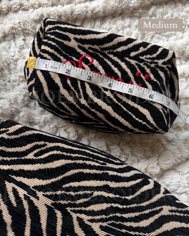 Puede incluir: Un estuche de tama&ntilde;o mediano con estampado de cebra en negro y beige. El estuche presenta texto bordado en rojo y se mide con una cinta m&eacute;trica blanca que muestra una longitud de aproximadamente 17,8 cm sobre la superficie superior.