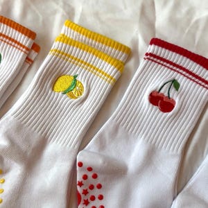 May include: White socks with embroidered fruit designs. One pair features a lemon, another cherries, and a third with an orange design. Each pair has colored stripes at the top and red non-slip grips on the soles.