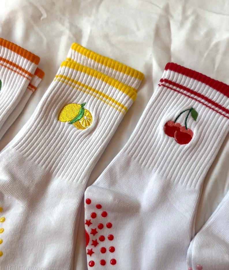 May include: White socks with embroidered fruit designs. One pair features a lemon, another cherries, and a third with an orange design. Each pair has colored stripes at the top and red non-slip grips on the soles.