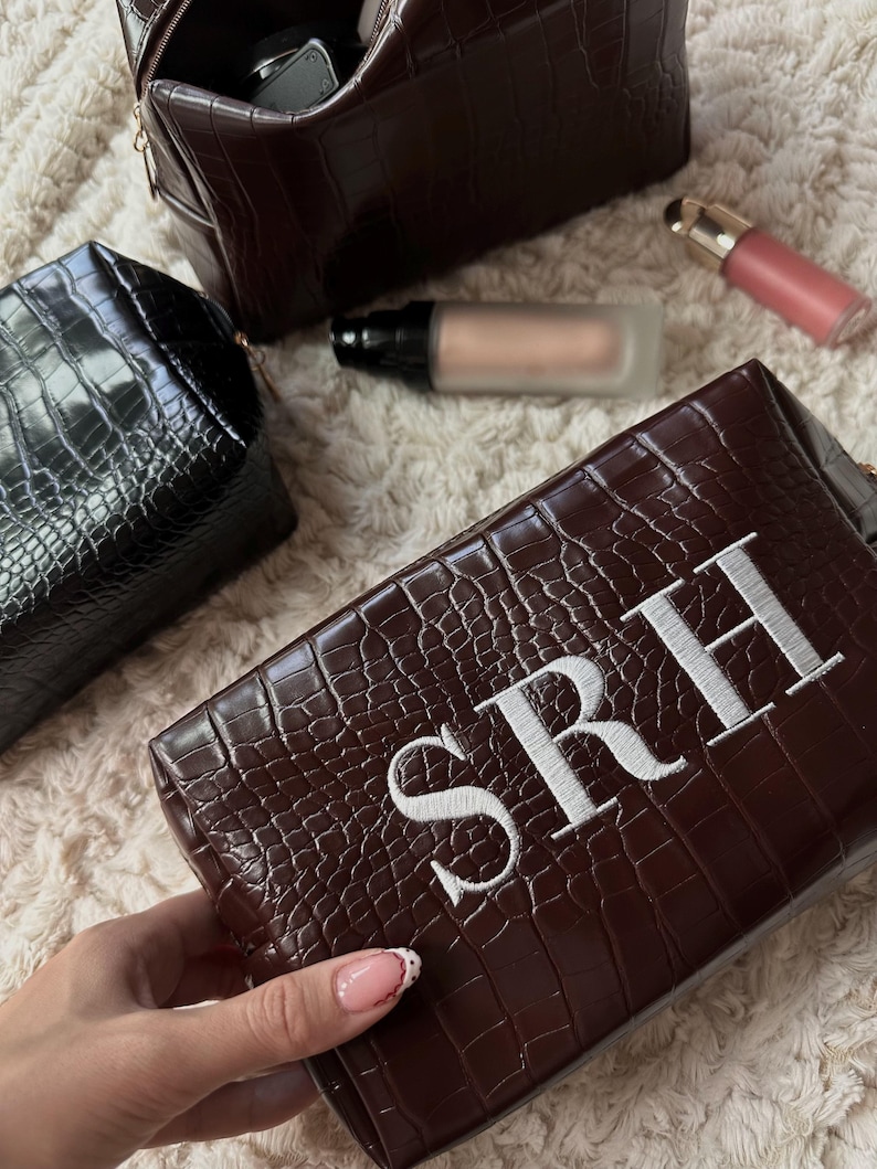 May include: Brown faux-leather cosmetic bag with white embroidered initials "SRH". The bag has a crocodile skin texture. A black cosmetic bag and a brown cosmetic bag are also visible, along with makeup products.
