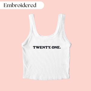 Pu&ograve; includere: Canotta bianca a coste con la scritta "TWENTY. ONE." in nero. La parola "Embroidered" &egrave; sopra la canotta. La canotta &egrave; su uno sfondo rosa chiaro.