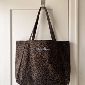 Op de afbeelding: Een bruine en zwarte tote bag met luipaardprint, voorzien van twee schouderbanden en de geborduurde witte tekst Mrs. Reyes in het midden. De stoffen tas heeft een rechthoekige vorm en hangt aan een zilverkleurige metalen haak.