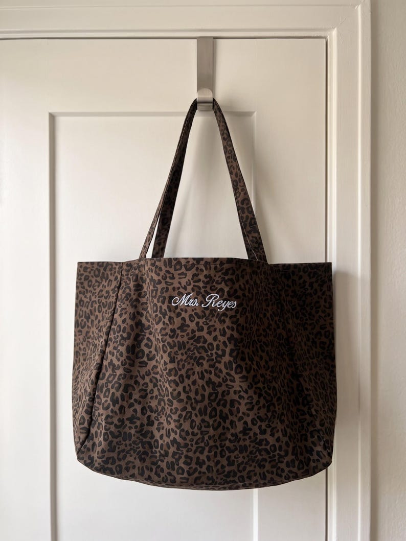 Op de afbeelding: Een bruine en zwarte tote bag met luipaardprint, voorzien van twee schouderbanden en de geborduurde witte tekst Mrs. Reyes in het midden. De stoffen tas heeft een rechthoekige vorm en hangt aan een zilverkleurige metalen haak.