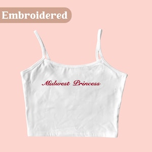 May include: White crop top with thin straps. The words "Midwest Princess" are embroidered in a cursive font across the front. The word "Embroidered" is in a light brown rectangle in the top left corner.