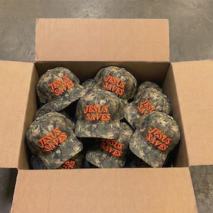 May include: A cardboard box filled with camouflage baseball caps. Each cap has the words "JESUS SAVES" embroidered in orange thread across the front. The caps are a mix of green, brown, and tan.