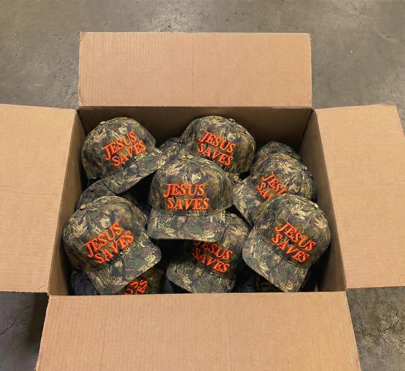 May include: A cardboard box filled with camouflage baseball caps. Each cap has the words "JESUS SAVES" embroidered in orange thread across the front. The caps are a mix of green, brown, and tan.