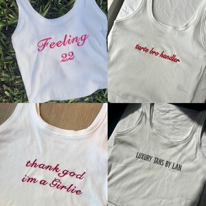 May include: Four white ribbed tank tops with embroidered text in pink and black. The phrases include "Feeling 22", "tarte bro handler", "thank god i'm a Girlie", and "LUXURY TANS BY LAN".