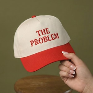 Pode incluir: Boné de beisebol com coroa creme e aba vermelha. As palavras "THE PROBLEM" são bordadas em vermelho na frente do boné. O boné é segurado por uma mão.