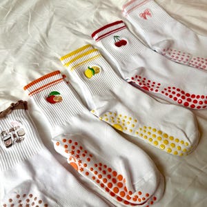 May include: White socks with colorful embroidered fruit designs, including a cherry, lemon, and orange. The socks have colored bands at the top and non-slip grips on the soles. The socks are designed for comfort and style.