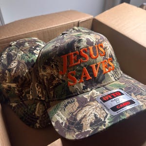 May include: Camouflage baseball cap with the text "JESUS SAVES" embroidered in orange thread. The cap is in a cardboard box. The cap has a curved brim and a classic fit.