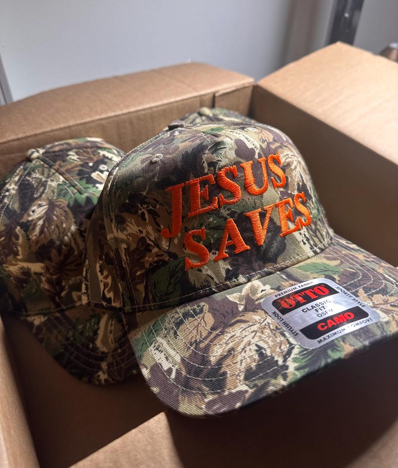 May include: Camouflage baseball cap with the text "JESUS SAVES" embroidered in orange thread. The cap is in a cardboard box. The cap has a curved brim and a classic fit.