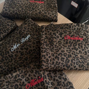 Op de afbeelding: Een collectie stoffen tasjes met een bruin en zwart luipaardpatroon, elk voorzien van gepersonaliseerd borduurwerk. De namen 'Mrs. Reyes', 'Christine' en 'Alicia' zijn met rood of wit garen op de voorzijde van de items geborduurd.