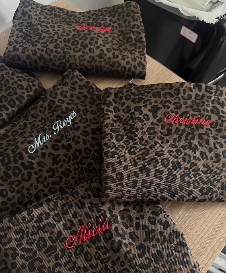 Op de afbeelding: Een collectie stoffen tasjes met een bruin en zwart luipaardpatroon, elk voorzien van gepersonaliseerd borduurwerk. De namen 'Mrs. Reyes', 'Christine' en 'Alicia' zijn met rood of wit garen op de voorzijde van de items geborduurd.