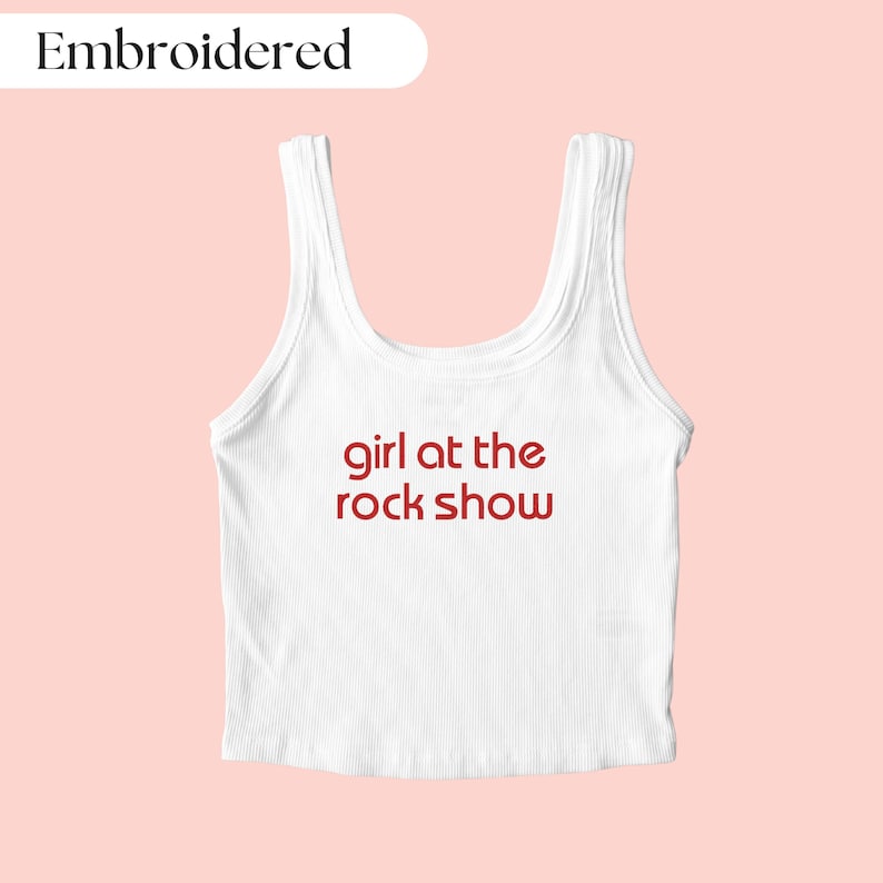 May include: White ribbed tank top with the text "girl at the rock show" in red. The top has a scoop neck and is sleeveless. The word "Embroidered" is in a white oval in the upper left corner.