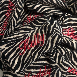 Puede incluir: Una colecci&oacute;n de peque&ntilde;as bolsas rectangulares de tela con un patr&oacute;n de rayas de cebra en negro y beige. Cada bolsa est&aacute; personalizada con un nombre bordado en hilo rojo en cursiva, incluyendo los nombres Kelly, Maddy, Courtney y Molly.