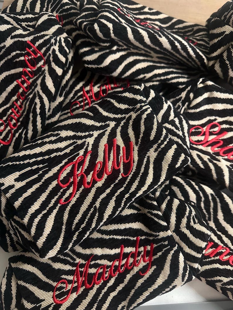 Puede incluir: Una colecci&oacute;n de peque&ntilde;as bolsas rectangulares de tela con un patr&oacute;n de rayas de cebra en negro y beige. Cada bolsa est&aacute; personalizada con un nombre bordado en hilo rojo en cursiva, incluyendo los nombres Kelly, Maddy, Courtney y Molly.