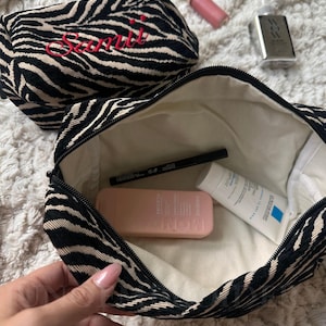 Puede incluir: Dos neceseres con cremallera y estampado de cebra en negro y beige. Uno est&aacute; cerrado y bordado con el texto rojo "Samii", mientras que el otro est&aacute; abierto, mostrando un interior color crema con un delineador negro, un frasco rosa y un tubo blanco.