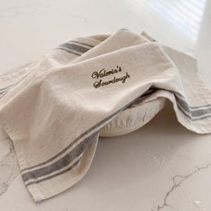 May include: A beige kitchen towel with dark gray stripes draped over a round proofing basket. The towel features dark green embroidered text that reads Valeria's Sourdough in a cursive script, providing a functional cover for bread dough.