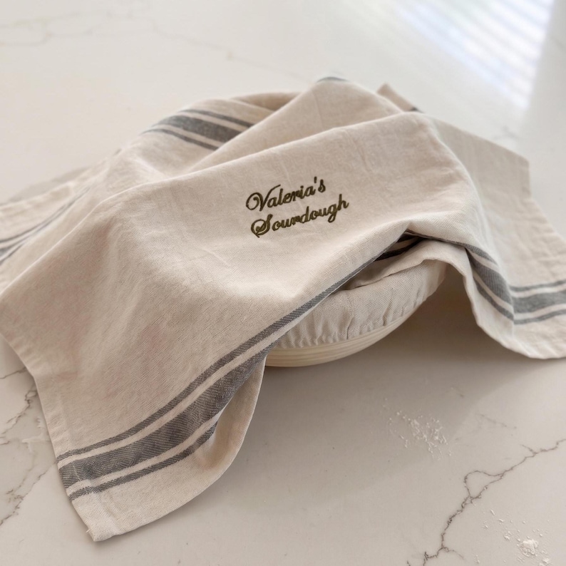 May include: A beige kitchen towel with dark gray stripes draped over a round proofing basket. The towel features dark green embroidered text that reads Valeria's Sourdough in a cursive script, providing a functional cover for bread dough.