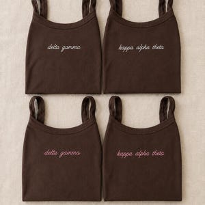 May include: Four dark brown tank tops arranged in a grid. The top left and bottom left tops feature the white and pink text delta gamma. The top right and bottom right tops feature the white and pink text kappa alpha theta in a cursive font.