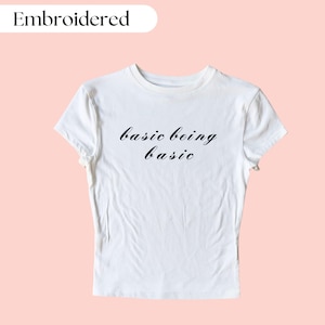 Könnte beinhalten: Weißes, kurzes T-Shirt mit dem in schwarzer Kursivschrift gestickten Text "basic being basic".