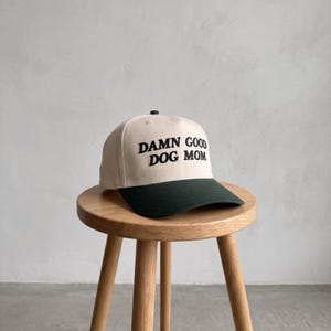 May include: A beige baseball cap with a dark green peak and the embroidered text "DAMN GOOD DOG MOM" in black. The cap is on a small wooden stool.