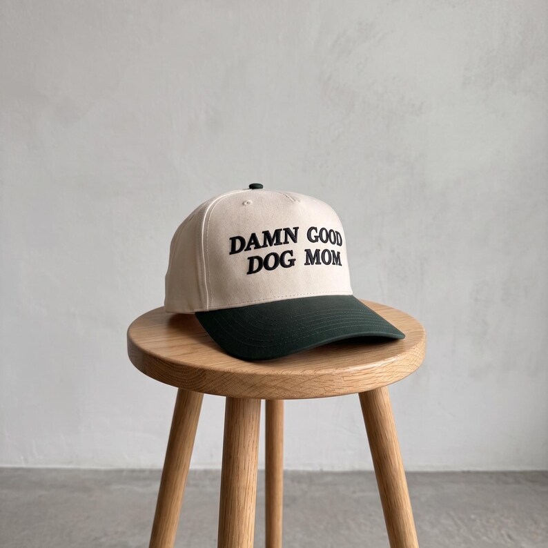 May include: A beige baseball cap with a dark green peak and the embroidered text "DAMN GOOD DOG MOM" in black. The cap is on a small wooden stool.