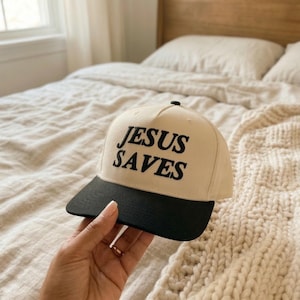 May include: A beige baseball cap with a black brim. The words "JESUS SAVES" are embroidered in black on the front of the cap. The cap is held by a person.