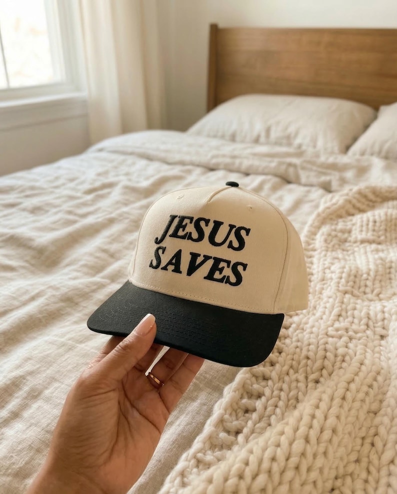 May include: A beige baseball cap with a black brim. The words "JESUS SAVES" are embroidered in black on the front of the cap. The cap is held by a person.