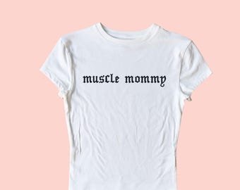 May include: White t-shirt with black embroidered text that reads "muscle mommy".