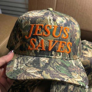 May include: Camouflage baseball cap with the embroidered text "JESUS SAVES" in orange. The cap features a curved brim and a button on top. The hat is a mix of green, brown, and tan colors.