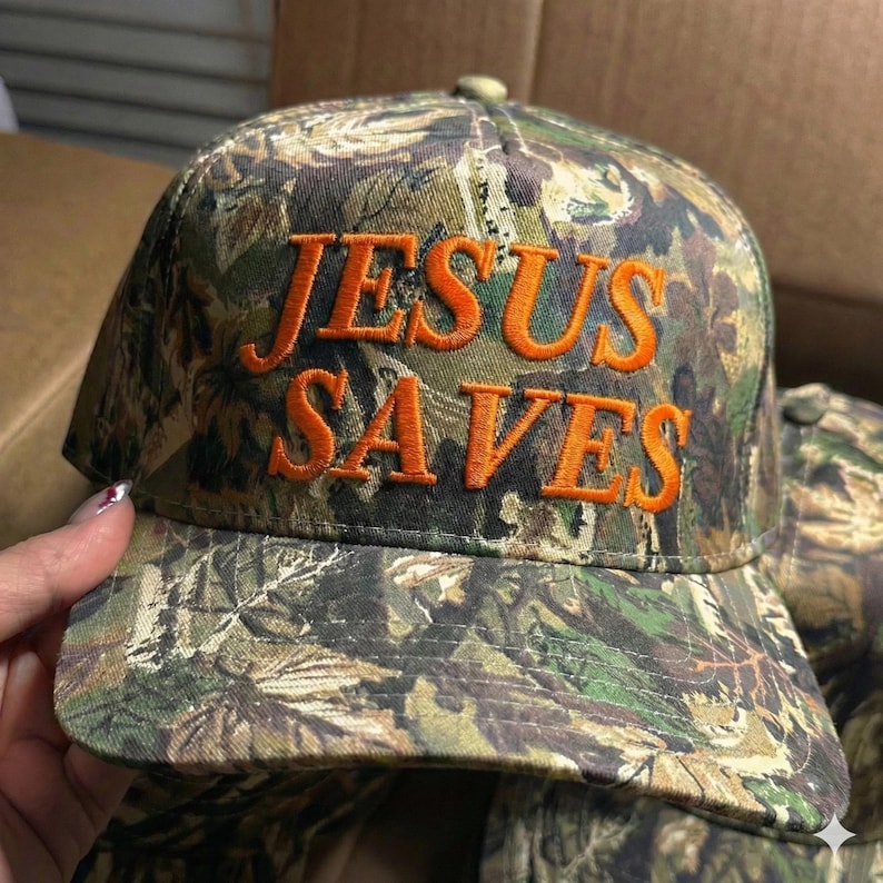 May include: Camouflage baseball cap with the embroidered text "JESUS SAVES" in orange. The cap features a curved brim and a button on top. The hat is a mix of green, brown, and tan colors.