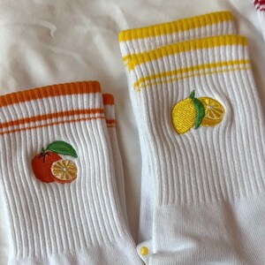 May include: White socks with orange and yellow striped cuffs. Each sock features an embroidered citrus fruit design, one with an orange and the other with a lemon. The socks are ribbed and have a casual style.