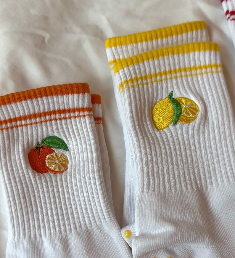 May include: White socks with orange and yellow striped cuffs. Each sock features an embroidered citrus fruit design, one with an orange and the other with a lemon. The socks are ribbed and have a casual style.