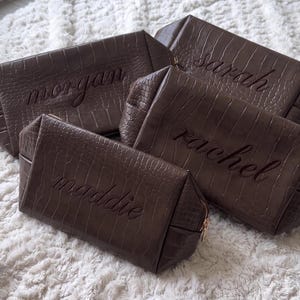 May include: Four dark brown rectangular toiletry bags with a textured crocodile skin pattern. Each bag features a gold-toned zipper and is personalized with an embroidered name in cursive script: morgan, sarah, rachel, and maddie.