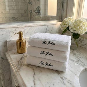 May include: Three white bath towels stacked on a marble countertop, each embroidered with "The Johns" in black script. A gold soap dispenser and a vase of white flowers are also visible.