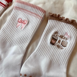 May include: White ribbed socks with pink accents and embroidered designs. One pair features a pink bow, while the other displays a coffee cup, pastry, and the words "Pilates & Coffee". The socks have brown non-slip grips on the soles.