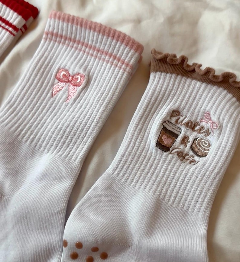 May include: White ribbed socks with pink accents and embroidered designs. One pair features a pink bow, while the other displays a coffee cup, pastry, and the words "Pilates & Coffee". The socks have brown non-slip grips on the soles.