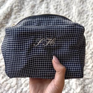 Può includere: Un beauty case a quadri blu navy e bianco con cerniera dorata. La borsa presenta le iniziali ricamate "J H" in corsivo. La borsa è tenuta in una mano.